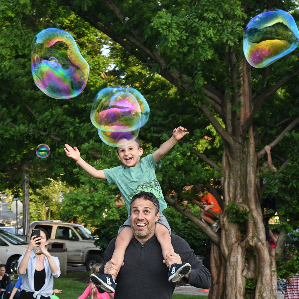 A child on a parent's shoulders chasing bubbles at Wicks Park
