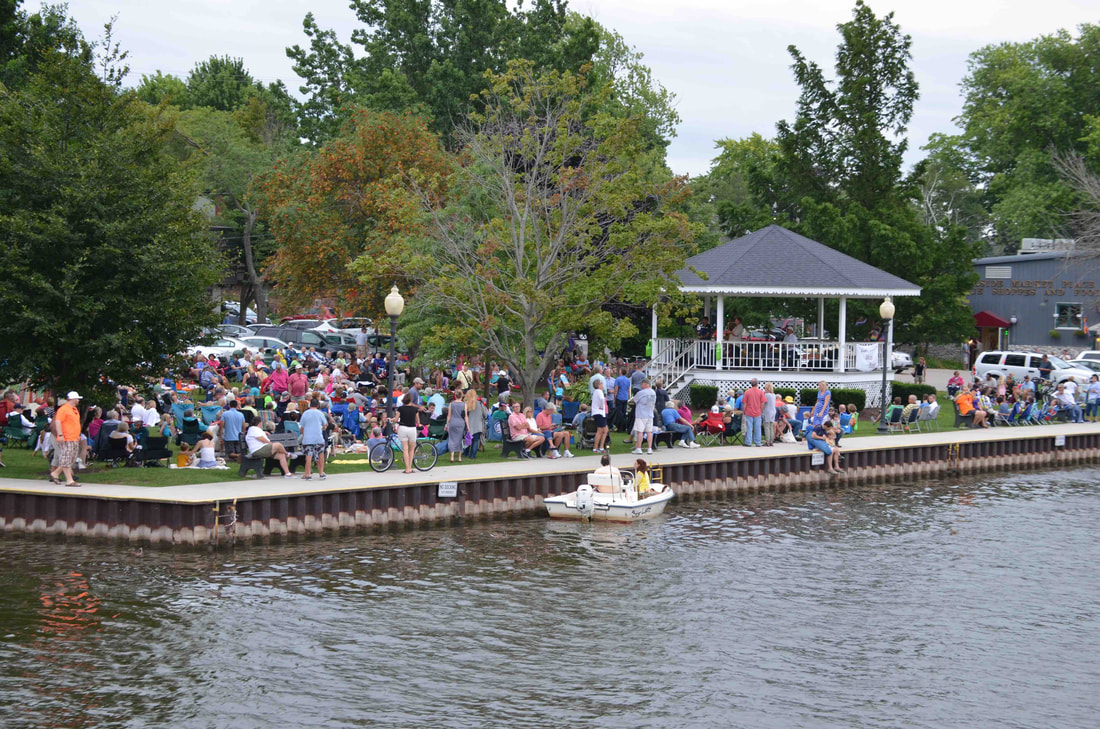 Boats and a crowd along the Kalamazoo River at Wicks Park on a summer evening